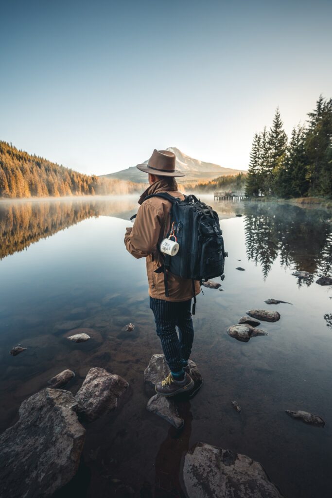 man in black jacket and brown hat standing on rock near lake 3217911